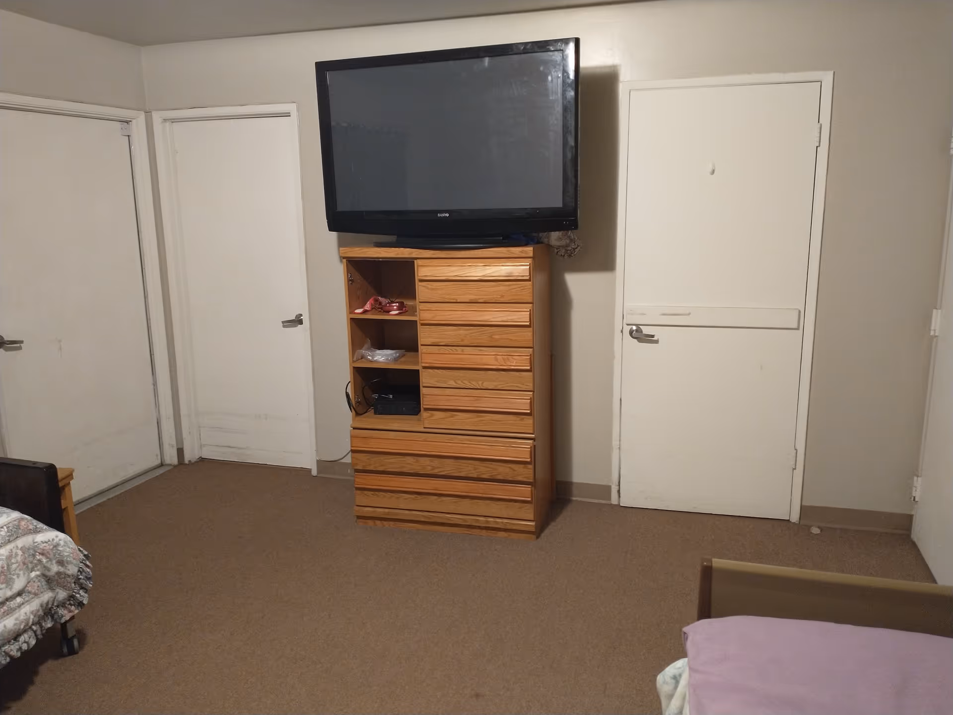 A simple bedroom with a wooden dresser holding a large flat-screen TV. There are three white doors on the walls, beige carpet, and part of a bed with a floral bedspread and a purple pillow visible.