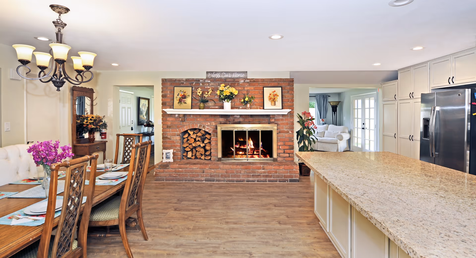 Open-concept interior showing a dining area and kitchen with a central brick fireplace and wood flooring.