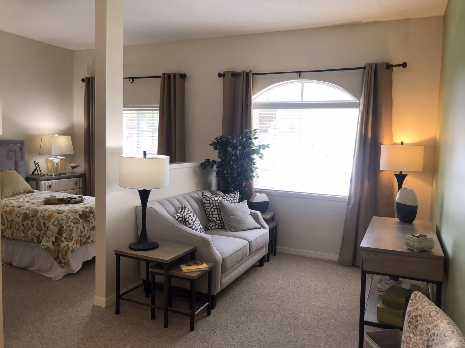 A cozy living area with a small gray loveseat adorned with patterned pillows, two side tables with lamps, and a large window with brown curtains letting in natural light. To the left, a partial view of a bedroom with a bed covered in a floral bedspread and a nightstand with a lamp and decorative items is visible.