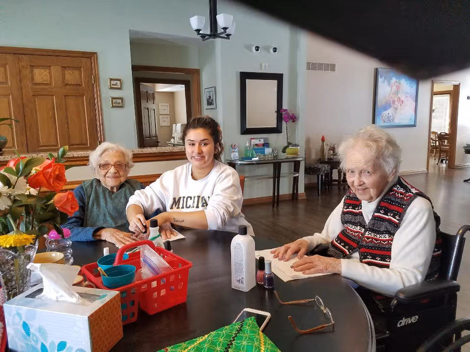 Three women seated around a table in a communal room doing nail care, with flowers and manicure supplies on the table.