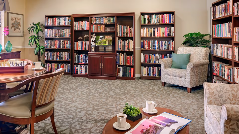 A cozy library room with multiple wooden bookshelves filled with books, two patterned armchairs with a green cushion on one, a round wooden table with two cups of coffee and an open magazine, a chessboard on another table, and some green plants and framed artwork on the walls.