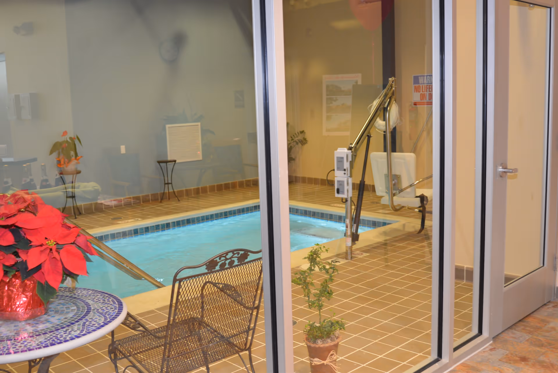 Indoor swimming pool area with a tiled floor, a metal chair and a round mosaic table with a red poinsettia plant on it. There is a pool lift for accessibility and some potted plants around the pool. The area is viewed through glass doors.