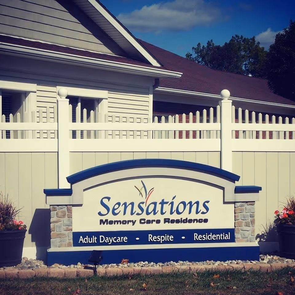 Outdoor sign for Sensations Memory Care Residence with a white fence and building in the background. The sign mentions services including Adult Daycare, Respite, and Residential care.