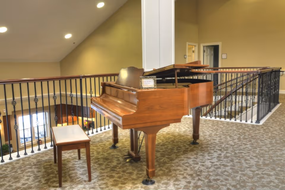 Grand piano and bench on a carpeted second-floor interior landing with decorative railing overlooking a lower level.