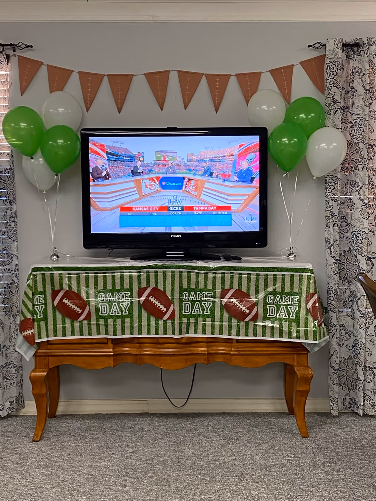 A television mounted on a wall displaying a sports broadcast with a football game theme. Below the TV is a wooden table covered with a green and white striped tablecloth decorated with footballs and the words 'GAME DAY'. On either side of the TV are clusters of green and white balloons. Above the TV is a brown pennant banner shaped like footballs. The room has patterned curtains on both sides of the TV.