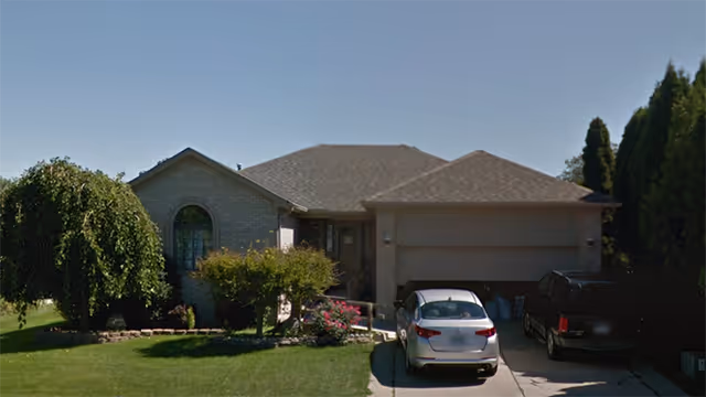 Front exterior view of a single-story residential house with a two-car garage, two cars parked in the driveway, and a well-maintained lawn with bushes and trees.