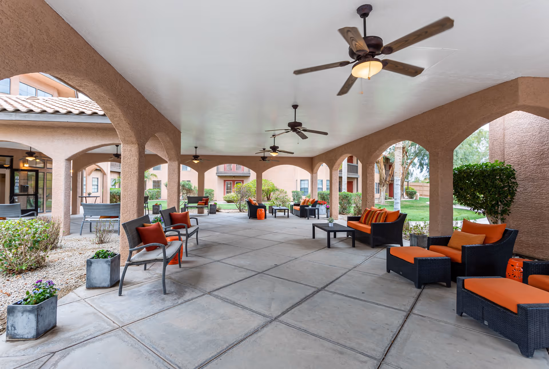 Covered outdoor seating area at Emerald Springs Senior Living with multiple ceiling fans, cushioned chairs and sofas with orange cushions, small tables, potted plants, and archways opening to a garden and building exterior.
