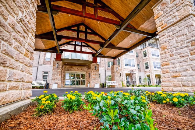 Entrance of a senior living facility with a covered driveway supported by stone pillars and a wooden ceiling. There are flower beds with yellow flowers and green plants in the foreground, and the building has multiple windows and balconies.
