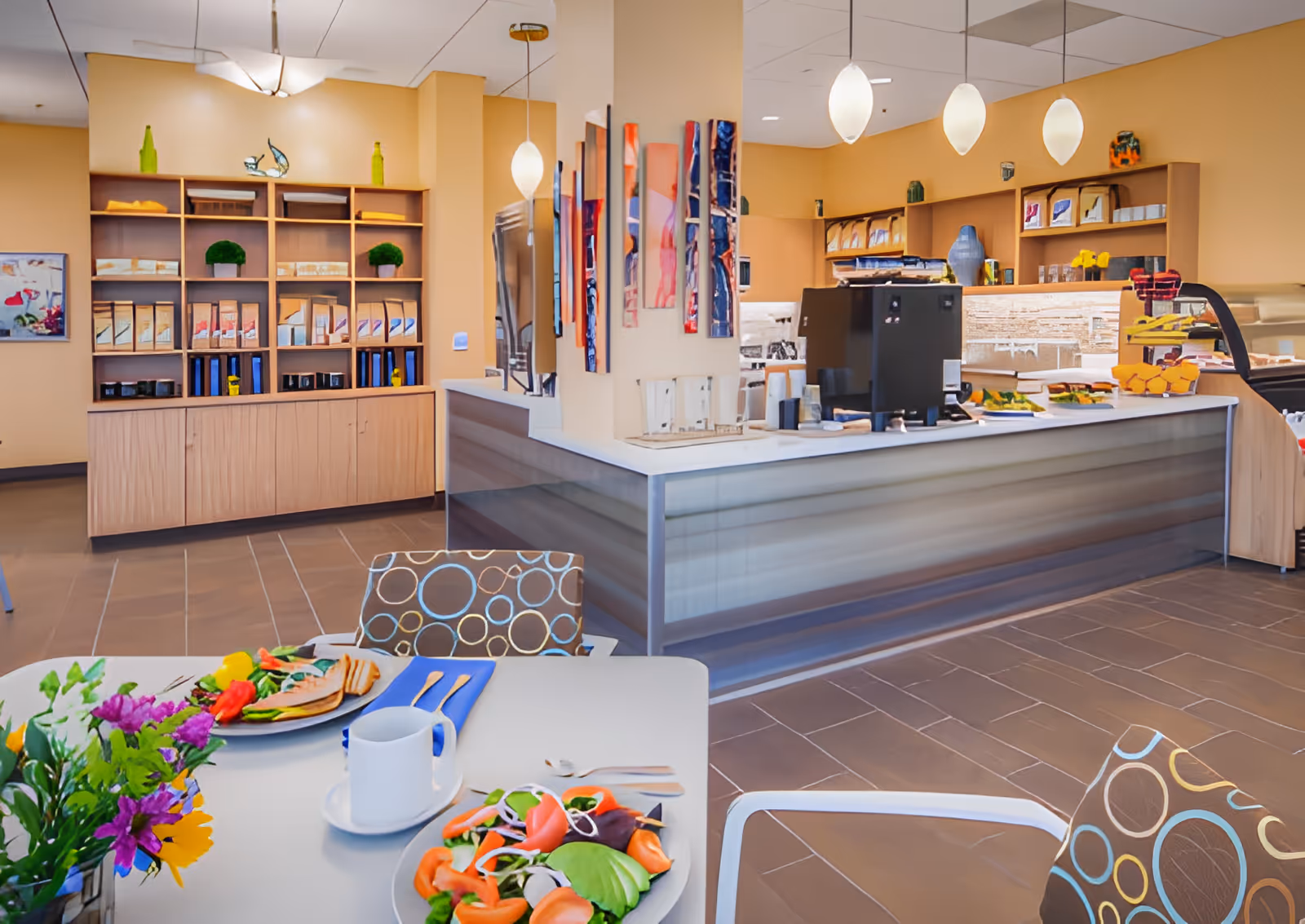 Bright senior facility dining area with set tables, patterned chairs, a service counter and shelving stocked behind the counter.