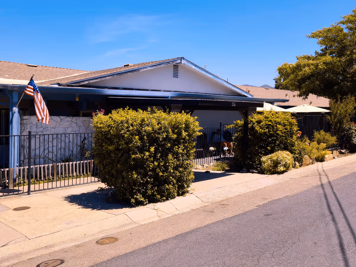 Exterior view of Country Haven Assisted Living facility showing a single-story building with a sloped roof, an American flag mounted on a blue post, and a black metal fence with bushes and greenery along the sidewalk under a clear blue sky.