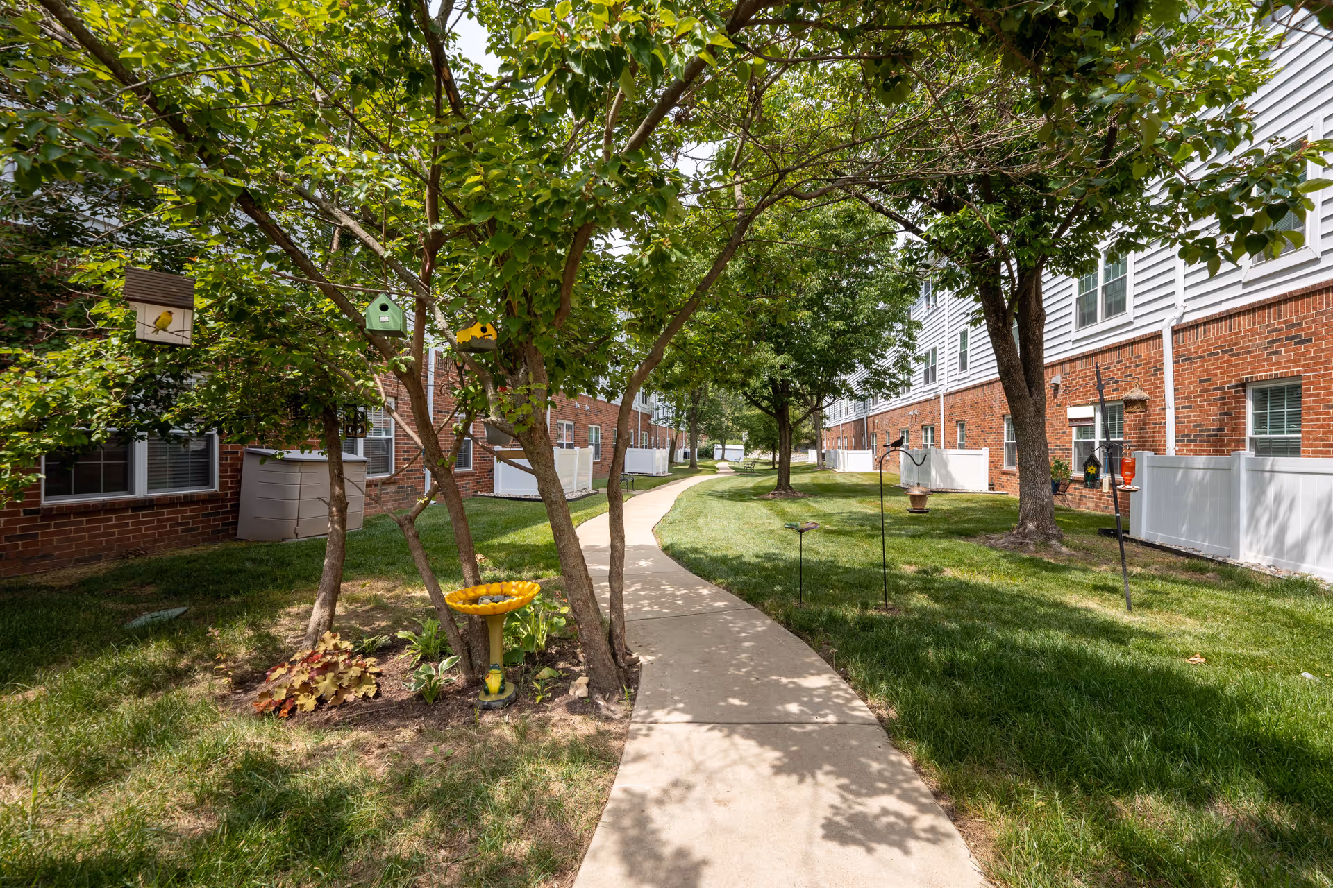 A paved walkway runs through a green outdoor area with grass, trees, and birdhouses hanging from the branches. The walkway is flanked by brick and white-paneled buildings with windows and white fences. There is a yellow birdbath and some small plants near the trees.