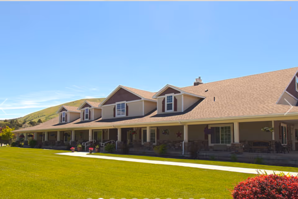 Exterior view of The Gables Assisted Living of Pocatello, showing a long building with a beige roof, multiple dormer windows, a covered porch with seating, and a well-maintained green lawn in front under a clear blue sky.