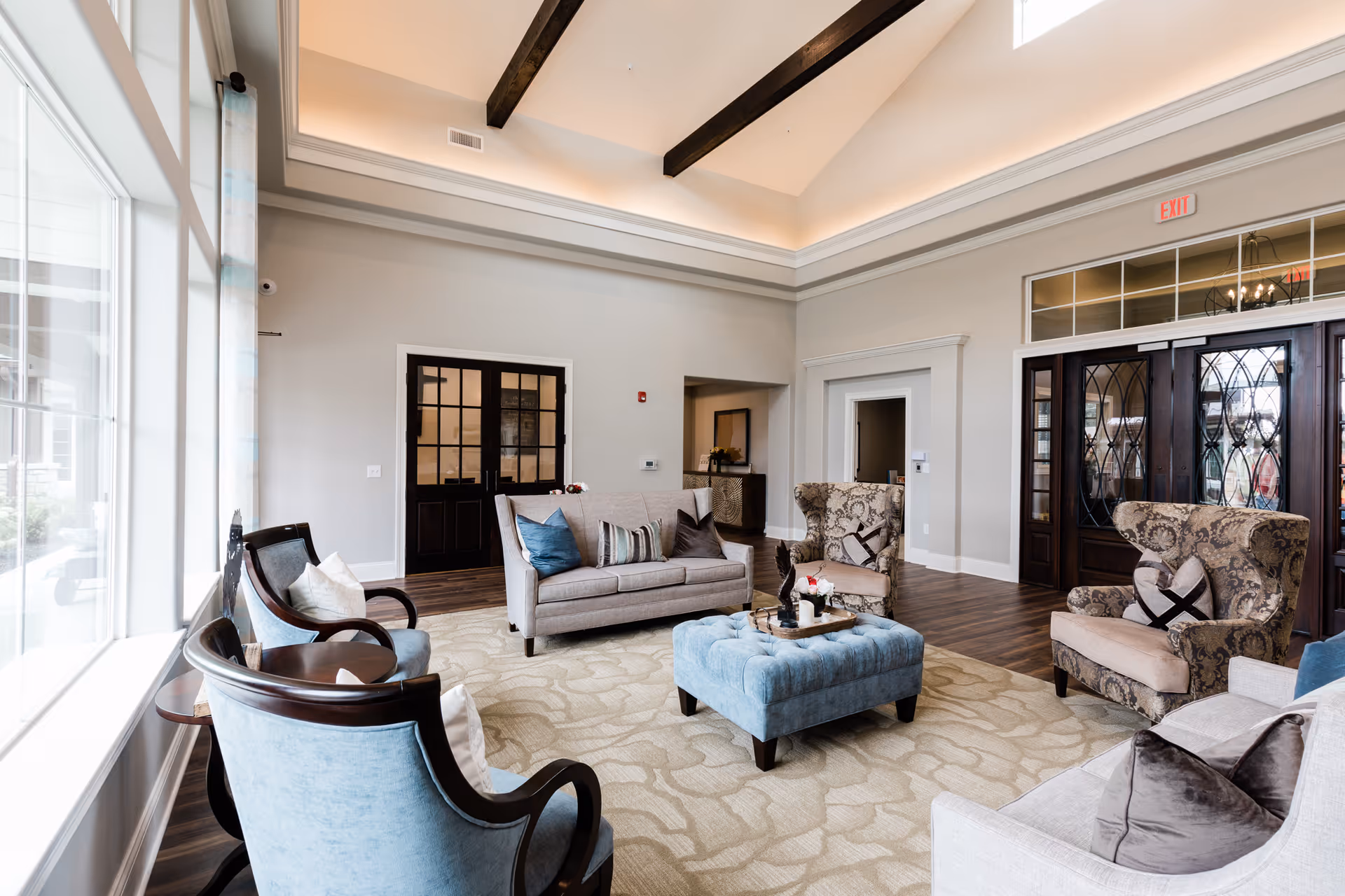 A bright and spacious living room with large windows on the left letting in natural light. The room features a beige patterned carpet, a light gray sofa with blue and striped pillows, two patterned armchairs, two blue upholstered chairs, and a blue tufted ottoman in the center with a tray holding decorative items. The ceiling is vaulted with exposed dark wooden beams and recessed lighting. There are dark wooden double doors with glass panels on the far wall and an exit sign above another set of doors.