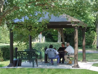Three people sitting and conversing under a gazebo in a green outdoor area with trees and grass.