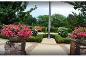 Outdoor garden area with a paved walkway leading to wooden benches surrounded by green bushes and trees. Large planters filled with vibrant pink flowers flank the walkway.