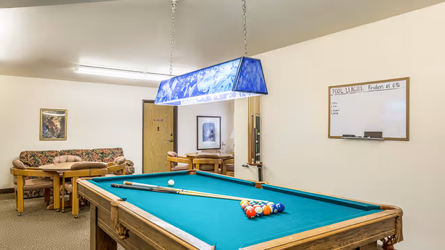 A recreation room with a pool table under a blue hanging light, a seating area with a couch and tables, and a whiteboard on the wall.