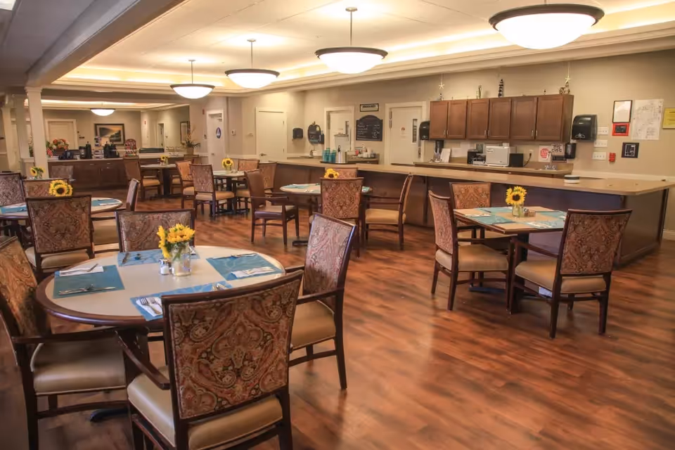 A communal dining room with round tables and upholstered chairs, place settings, and sunflower centerpieces.