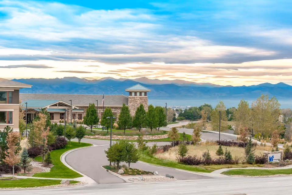 View of the Vi at Highlands Ranch senior living facility with landscaped grounds, trees, and a winding driveway. The building features stone and beige exterior walls with a tower-like structure. Mountains are visible in the background under a partly cloudy sky during sunset.