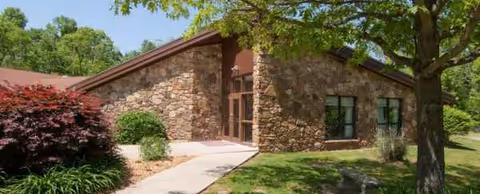 Stone exterior nursing facility entrance with glass doors, surrounded by trees and landscaping.