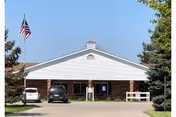 Front exterior view of Medilodge of Sterling building with a driveway, two parked cars, an American flag on a flagpole, and trees on either side under a clear blue sky.