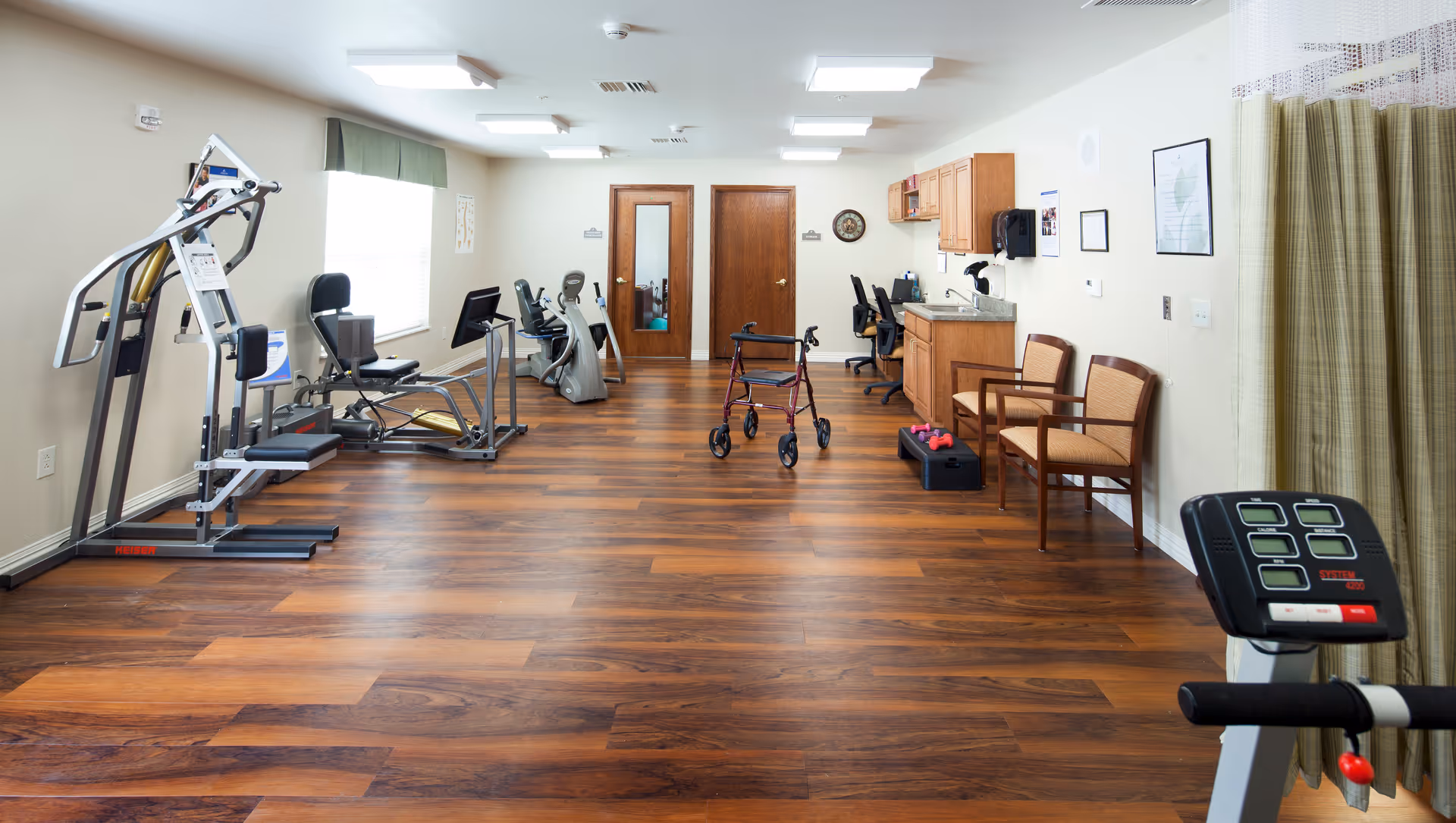 A senior living facility exercise room with various fitness equipment including a multi-station gym machine, stationary bikes, a walker, and small dumbbells. The room has wood flooring, white walls, a window with a green valance, two wooden doors, a small kitchenette area with a sink and cabinets, two chairs, and a curtain on the right side.