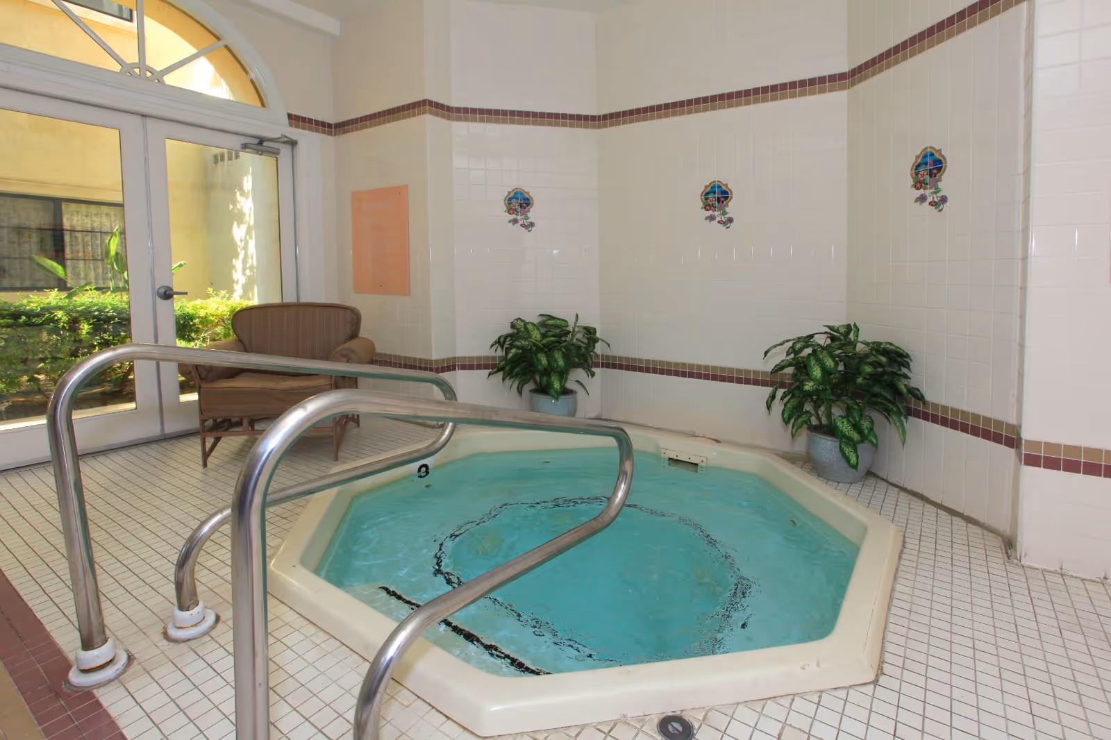 Indoor hot tub with clear blue water surrounded by white tiled walls and floor. There are two green potted plants on the tiled ledge behind the hot tub and a wicker chair near a glass door leading outside.