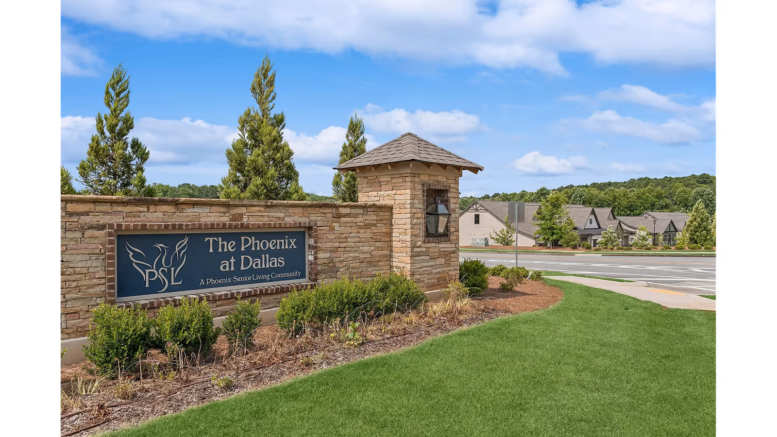 Stone entrance sign for The Phoenix at Dallas, a Phoenix Senior Living Community, surrounded by neatly trimmed bushes and green grass under a blue sky with some clouds. Residential buildings and trees are visible in the background.