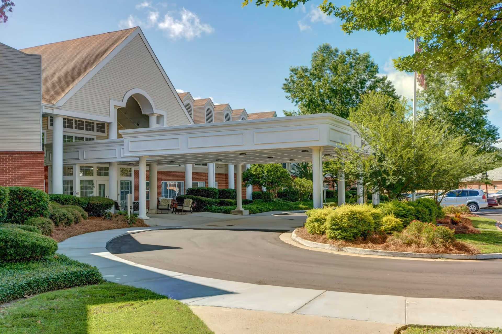 Exterior view of Ridgeland Place senior living facility entrance with a covered drop-off area, surrounded by well-maintained landscaping including bushes and trees under a blue sky.