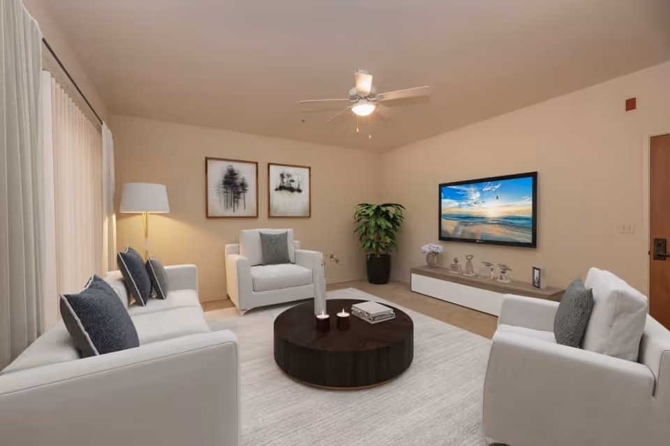 A modern senior living facility living room with beige walls, a ceiling fan, and a large window covered by light curtains. The room features a white sofa and two white armchairs with dark gray cushions, a round dark wooden coffee table with candles and books, a floor lamp, two framed black and white tree artworks on the wall, a potted plant, and a wall-mounted flat screen TV displaying a beach scene.