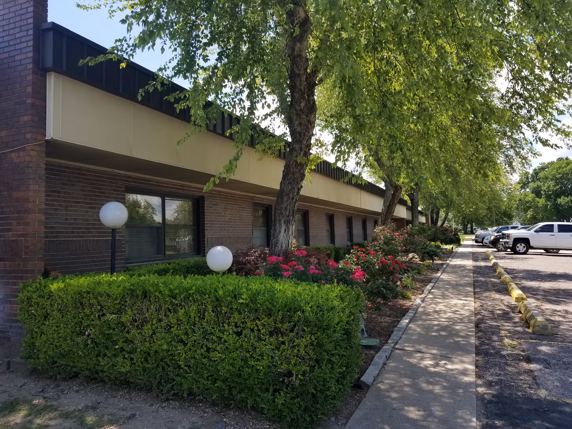 Low one-story brick nursing center front with trees, hedges, flowering bushes, a sidewalk and parked cars.