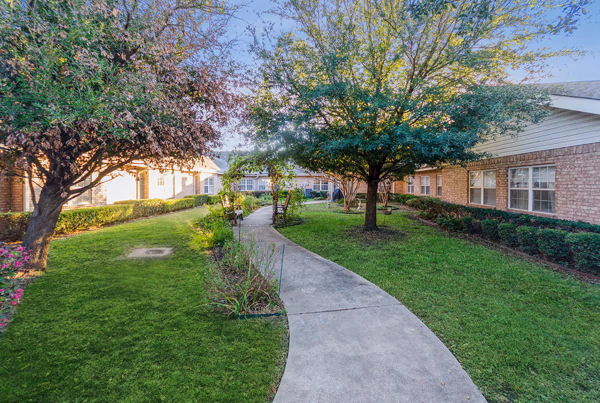 A curved concrete pathway runs through a green courtyard with well-maintained grass, bushes, and several trees. The courtyard is surrounded by single-story brick buildings with multiple windows. There are benches along the pathway under the trees, and the sky is clear and blue.