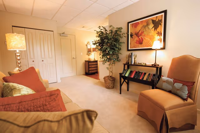 A cozy living room area with a beige sofa adorned with red and green pillows on the left, an orange upholstered chair with pillows on the right, a small black bookshelf with books and a lamp, a large framed artwork of autumn leaves on the wall, a tall potted plant, and a side table with a lamp near a closed door and closet.
