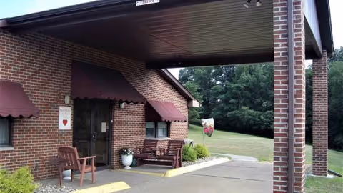 Brick facility entrance with maroon awnings, benches, and a grassy, tree-filled exterior.