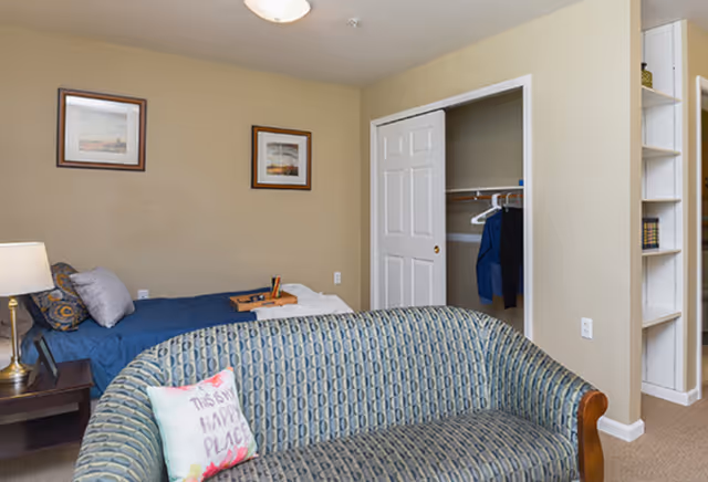 A cozy senior living bedroom with a single bed covered in blue bedding, a wooden nightstand with a lamp, and two framed pictures on the beige wall. In the foreground, there is a patterned couch with a decorative pillow that reads 'This is my happy place.' An open closet with clothes hanging inside and a built-in bookshelf are visible on the right side.