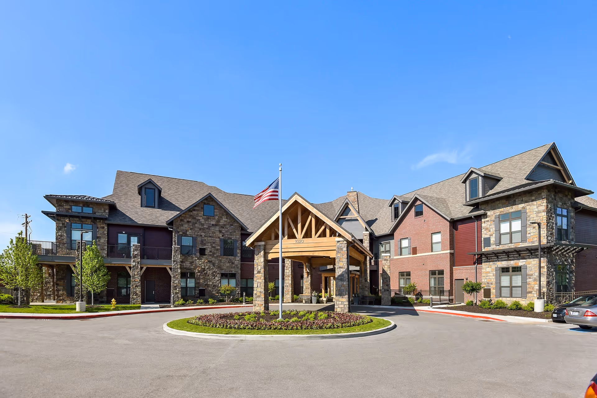 Front exterior view of the American House Town and Country senior living facility with a circular driveway, landscaped greenery, and an American flag on a flagpole under a wooden entrance canopy.