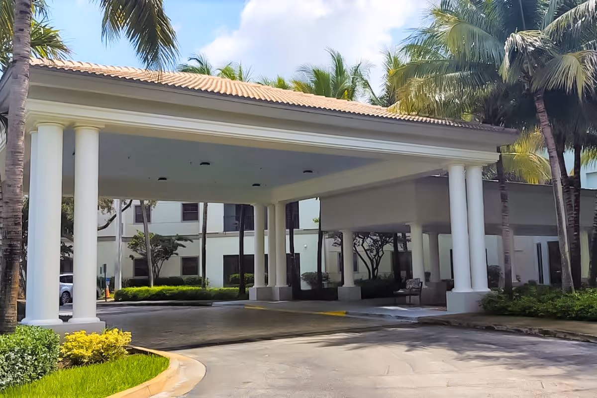 Entrance area of a senior living facility with a covered driveway supported by white columns, surrounded by palm trees and greenery under a partly cloudy sky.
