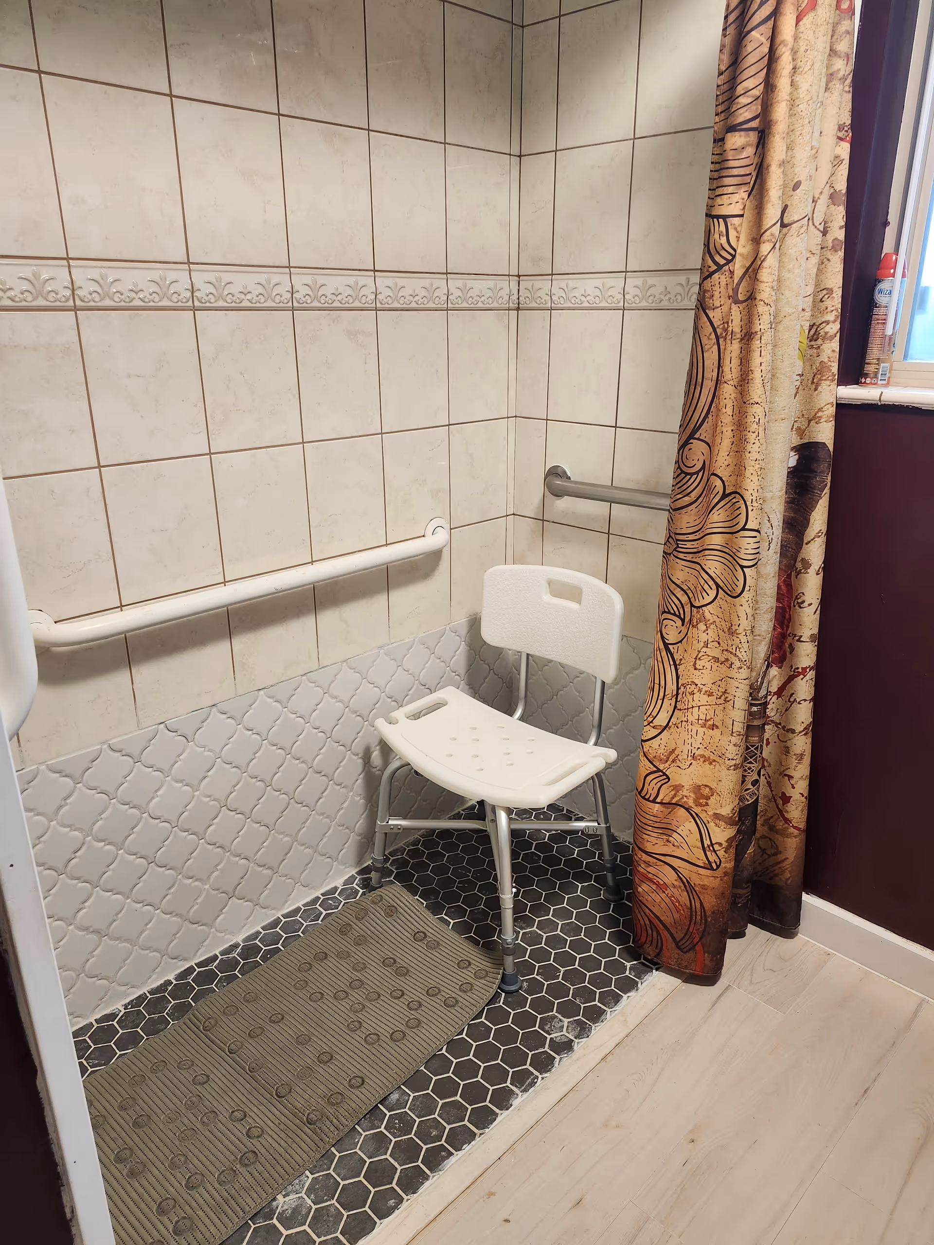 A tiled shower area with a white plastic shower chair, grab bars on the walls, a textured floor mat, and a decorative shower curtain partially drawn to the side. The walls have white and light gray tiles, and there is a window with a can of spray visible on the windowsill.