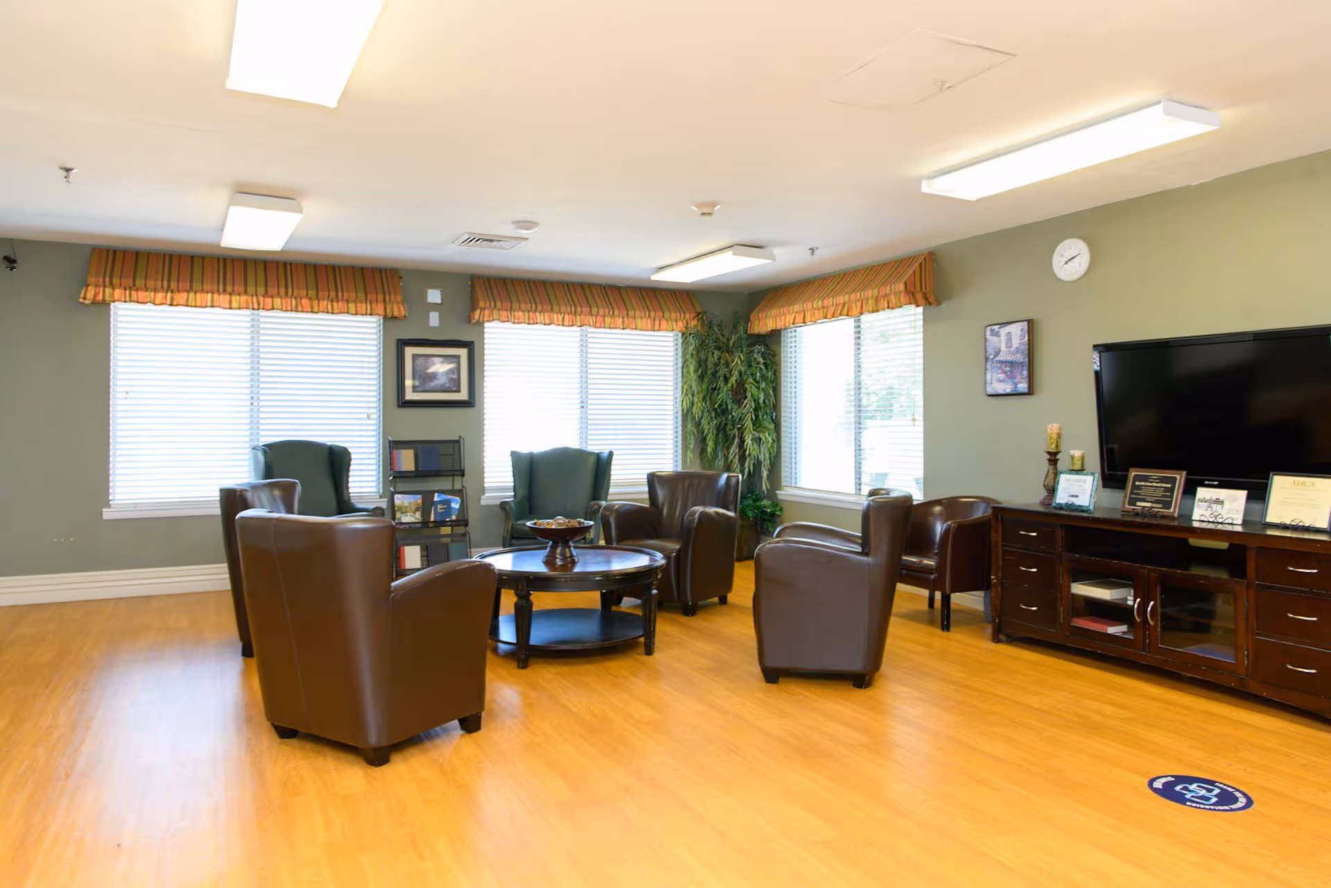 A well-lit living room with wooden flooring, several brown leather armchairs arranged around a round coffee table, two green wingback chairs near the windows, a large flat-screen TV on a dark wooden cabinet, and large windows with striped valances allowing natural light to fill the room.