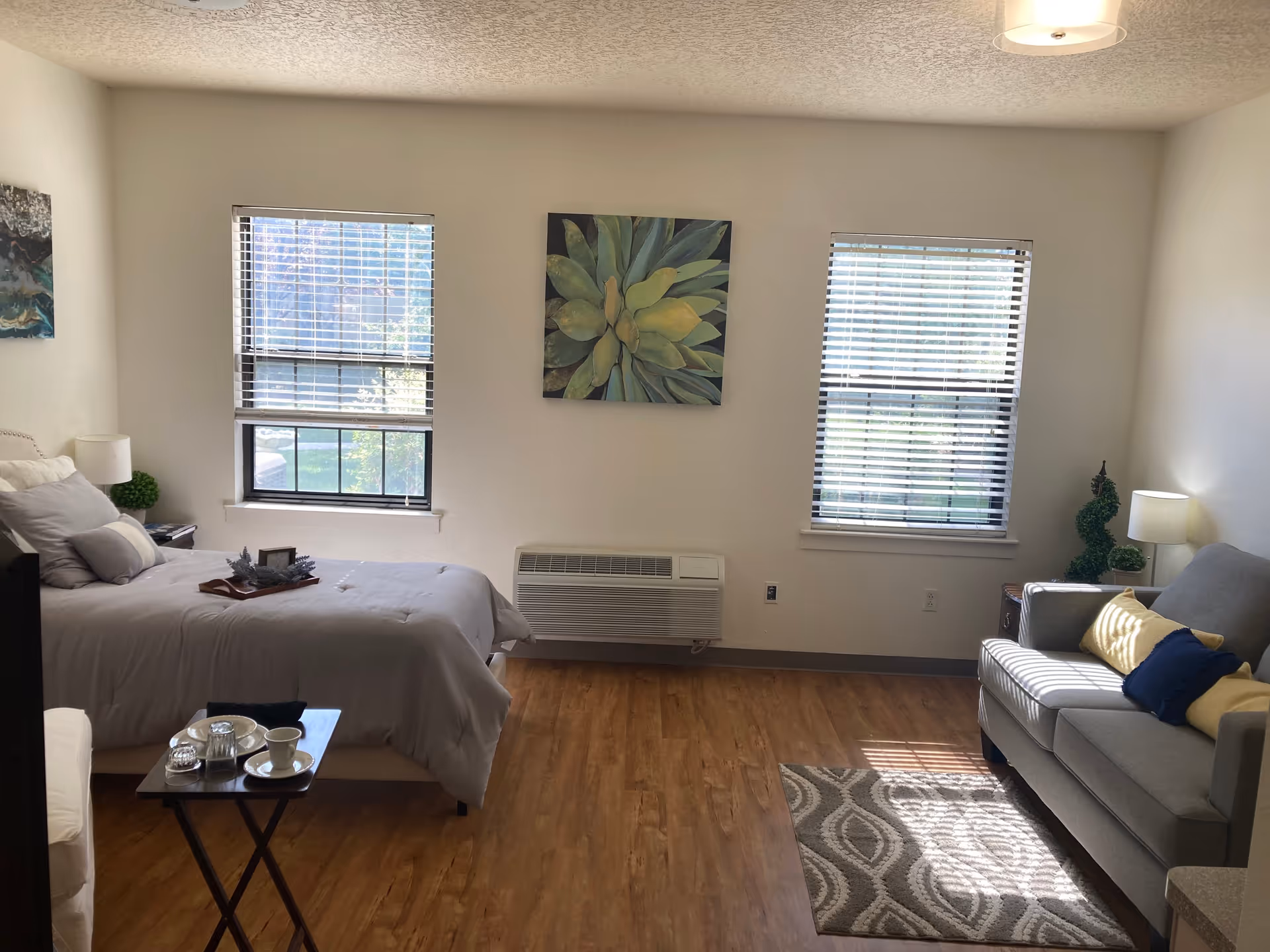 A bright and cozy bedroom with two windows letting in natural light. The room features a neatly made bed with gray bedding and pillows, a small side table with a lamp and plant, a gray sofa with yellow and blue cushions, a patterned rug, and a small table with a tea set. There is a green plant decoration near the sofa and a large painting of green leaves on the wall between the windows.