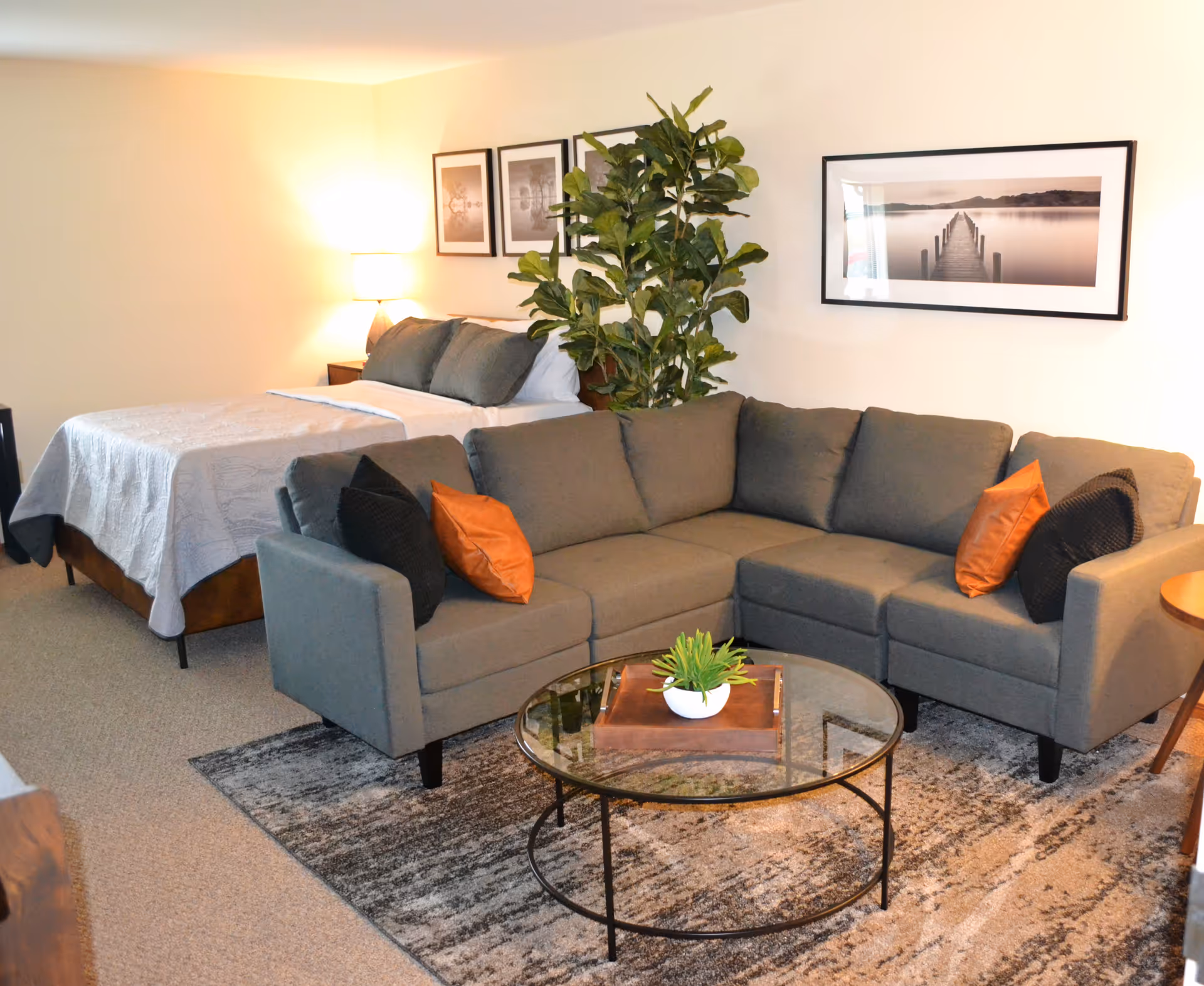 A cozy living area with a gray sectional sofa adorned with orange and black pillows, a round glass coffee table with a small plant on a wooden tray, and a bed with white bedding and gray pillows in the background. The room features framed black and white photos on the walls and a tall green plant beside the bed.