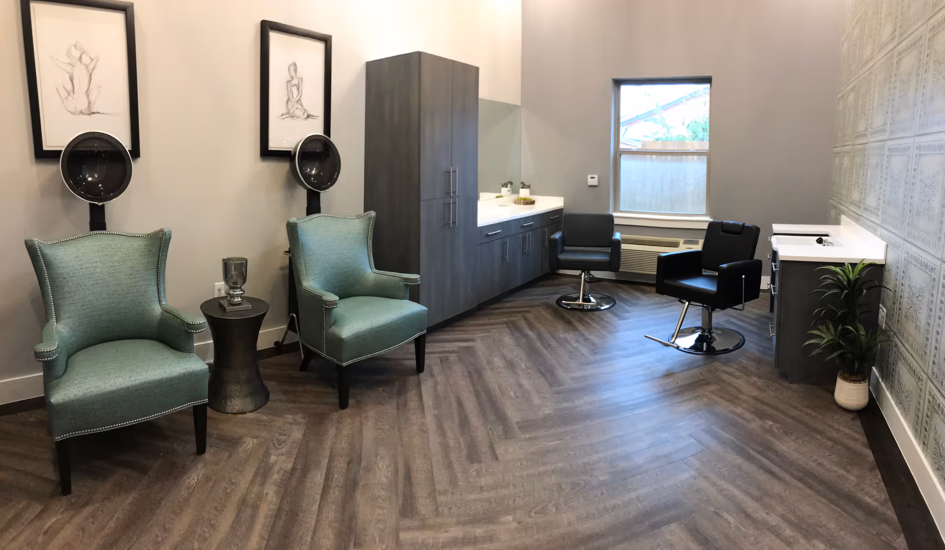 A modern salon room with two teal armchairs under hair dryers on the left, two black salon chairs on the right, a long cabinet with a sink in the back, a window, and a potted plant in the corner. The floor has a wood pattern and the walls are painted gray with framed artwork and decorative wallpaper.