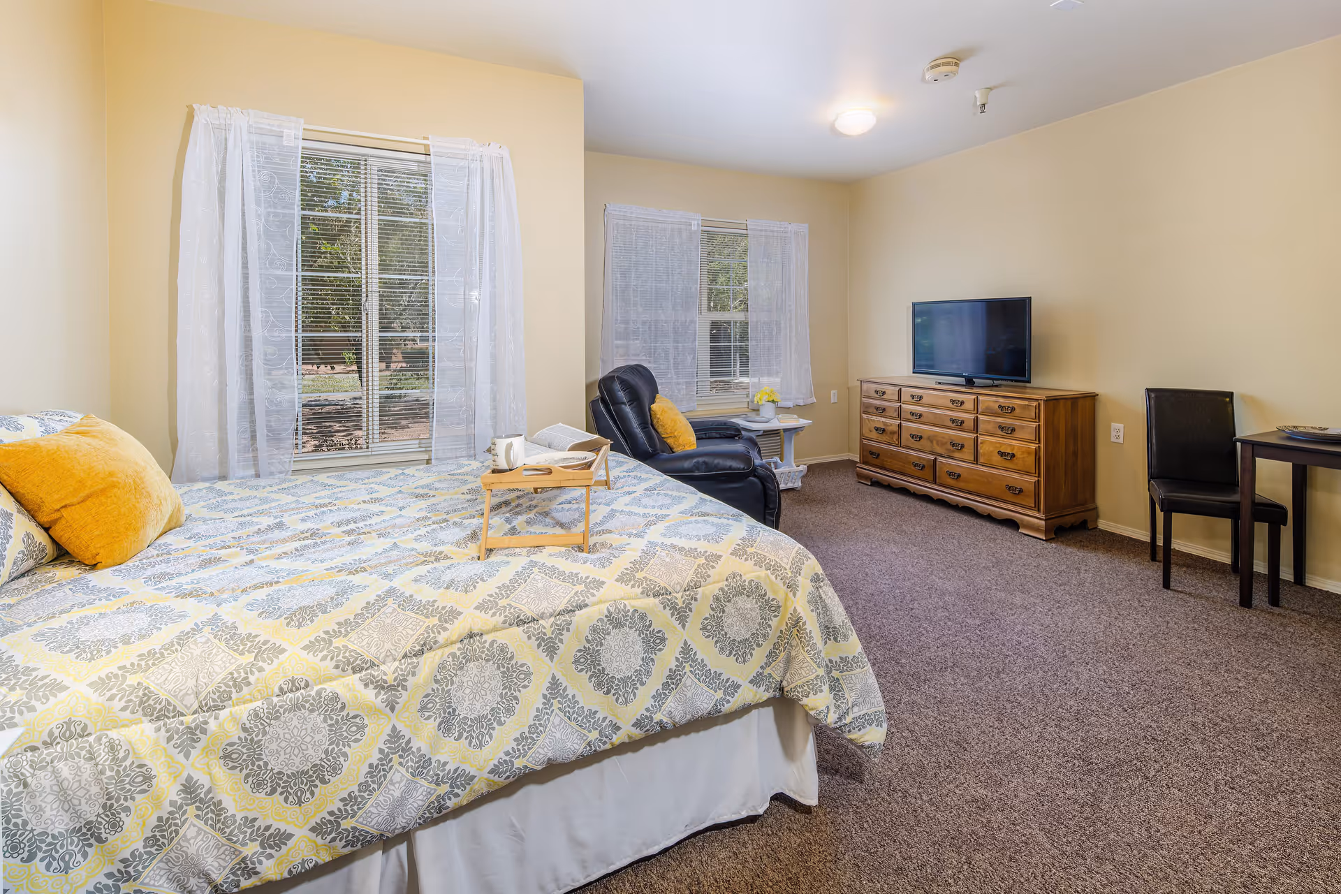 A bright bedroom with a large bed covered in a patterned yellow and gray bedspread, two yellow pillows, and a wooden tray with a cup and book. The room has two windows with sheer white curtains, a black recliner chair with a yellow pillow, a wooden dresser with a flat-screen TV on top, and a small table with a black chair. The walls are painted light yellow and the floor is carpeted.