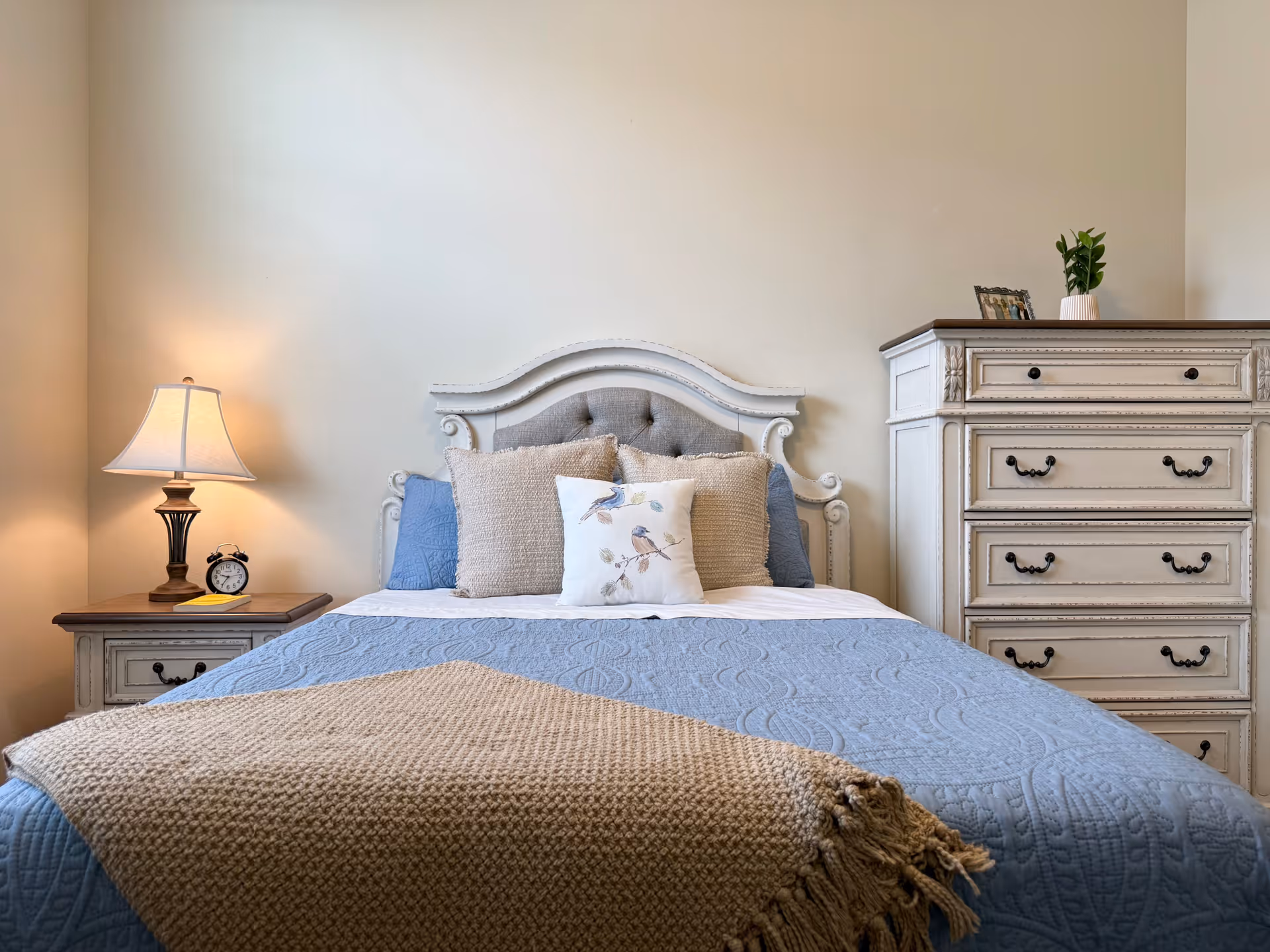 A neatly made bed with a blue quilt and beige throw blanket in a senior living bedroom. The bed has a decorative headboard and several pillows, including one with a bird design. To the left is a nightstand with a lamp and an alarm clock, and to the right is a white dresser with decorative handles and a small plant on top.