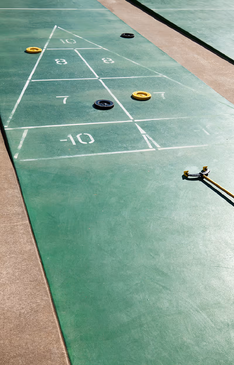 Outdoor shuffleboard court with green playing surface, white markings, and several yellow and black shuffleboard discs placed on the court.