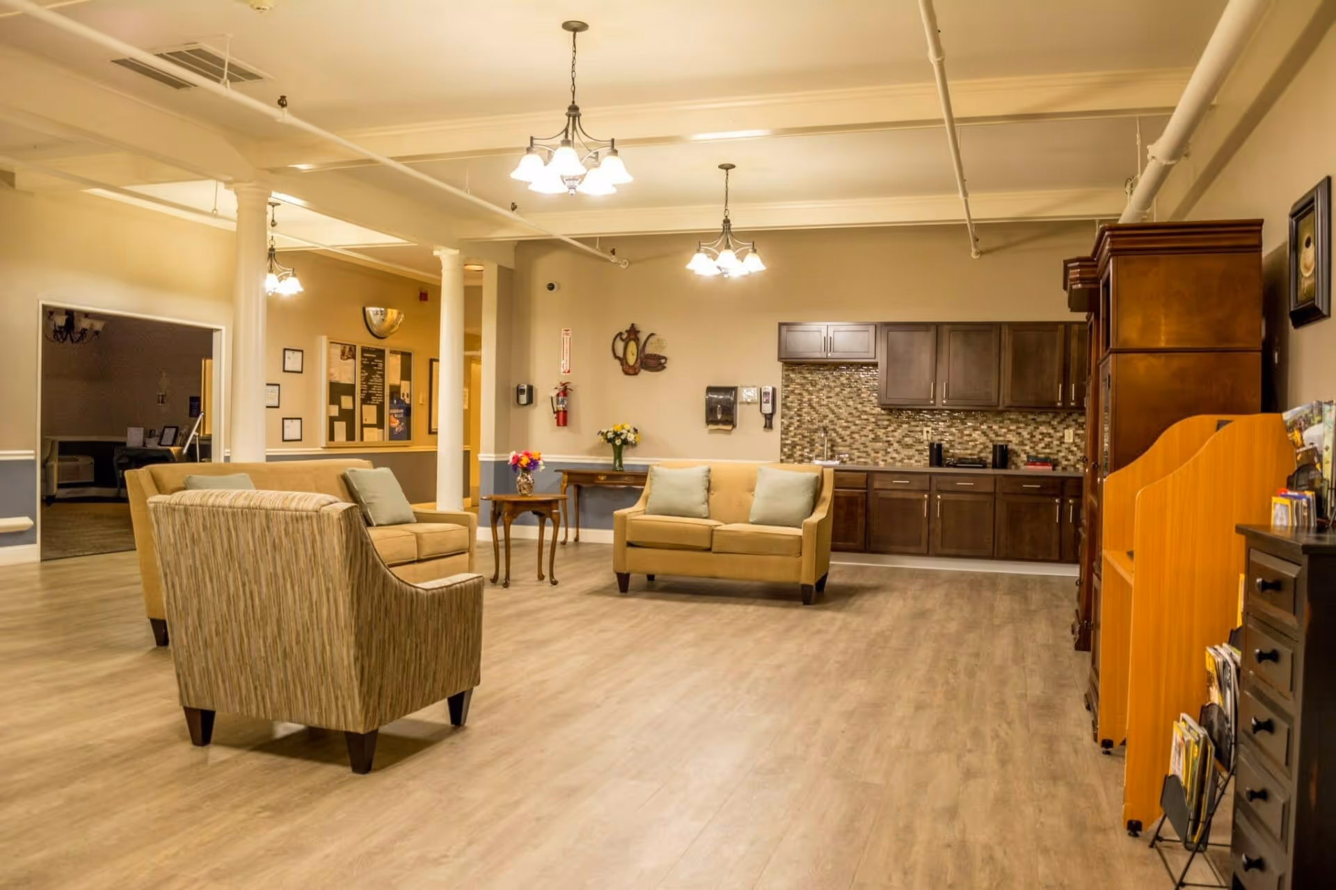 A spacious communal lounge with sofas, armchairs, side tables, and a kitchenette along the back wall.