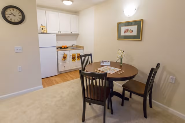 A small dining area with a round wooden table and three chairs, adjacent to a compact kitchen with white cabinets, a refrigerator, and a sink. The walls are light-colored, and a framed picture hangs above the table. A clock is mounted on the wall near the kitchen.