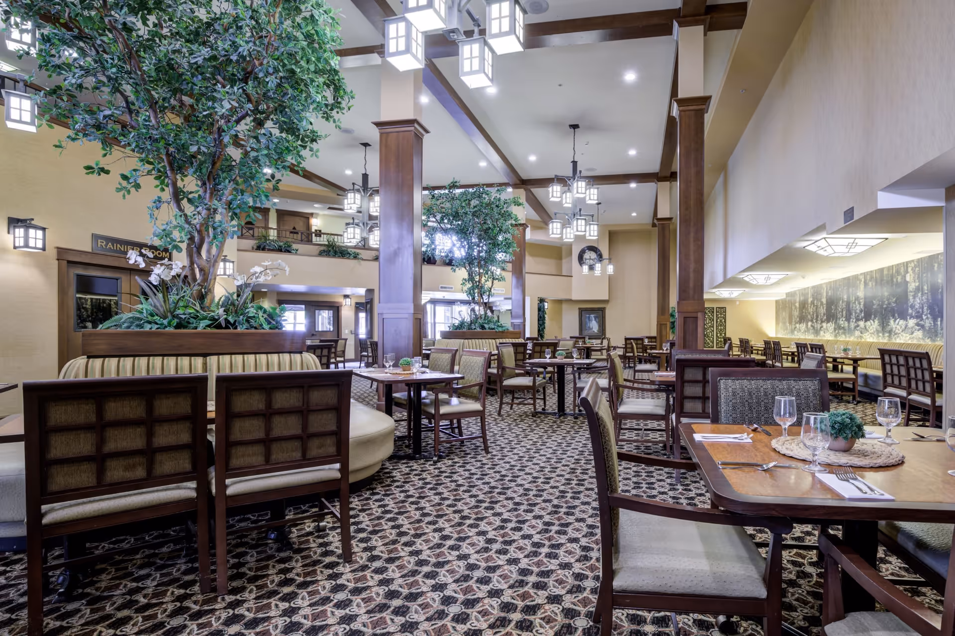 Spacious dining room with multiple wooden tables and chairs arranged neatly. The room features patterned carpet flooring, large potted plants, and warm lighting from hanging lantern-style fixtures. There are booths along the walls and a high ceiling with exposed wooden beams.