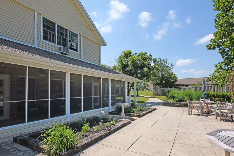Outdoor patio area at Wellington Manor Knoxville featuring a paved walkway, garden beds with plants, a birdbath, and several wooden tables and chairs with umbrellas under a clear blue sky.