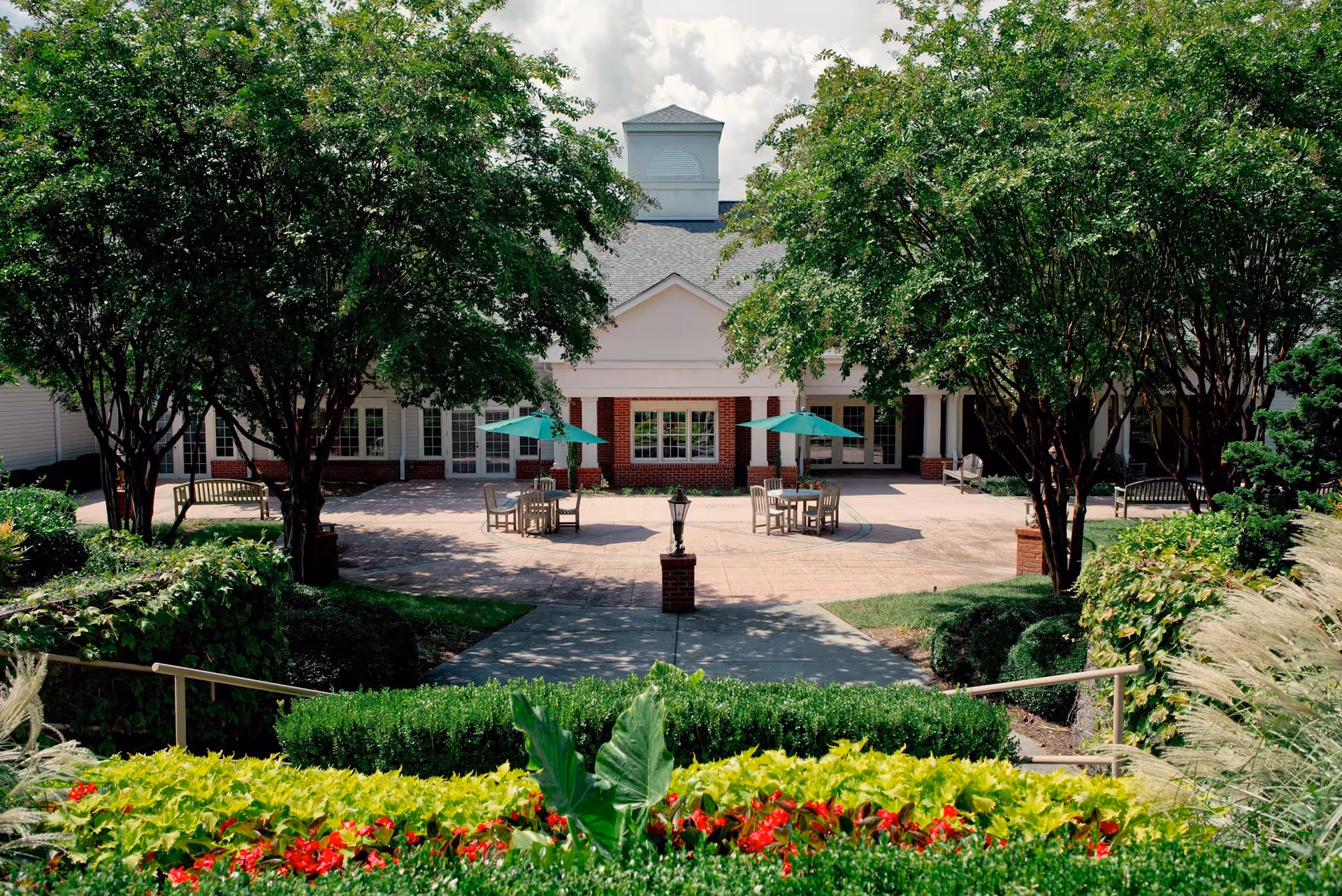 A landscaped outdoor courtyard with tables, umbrellas and benches in front of a brick building, framed by trees and flower beds.