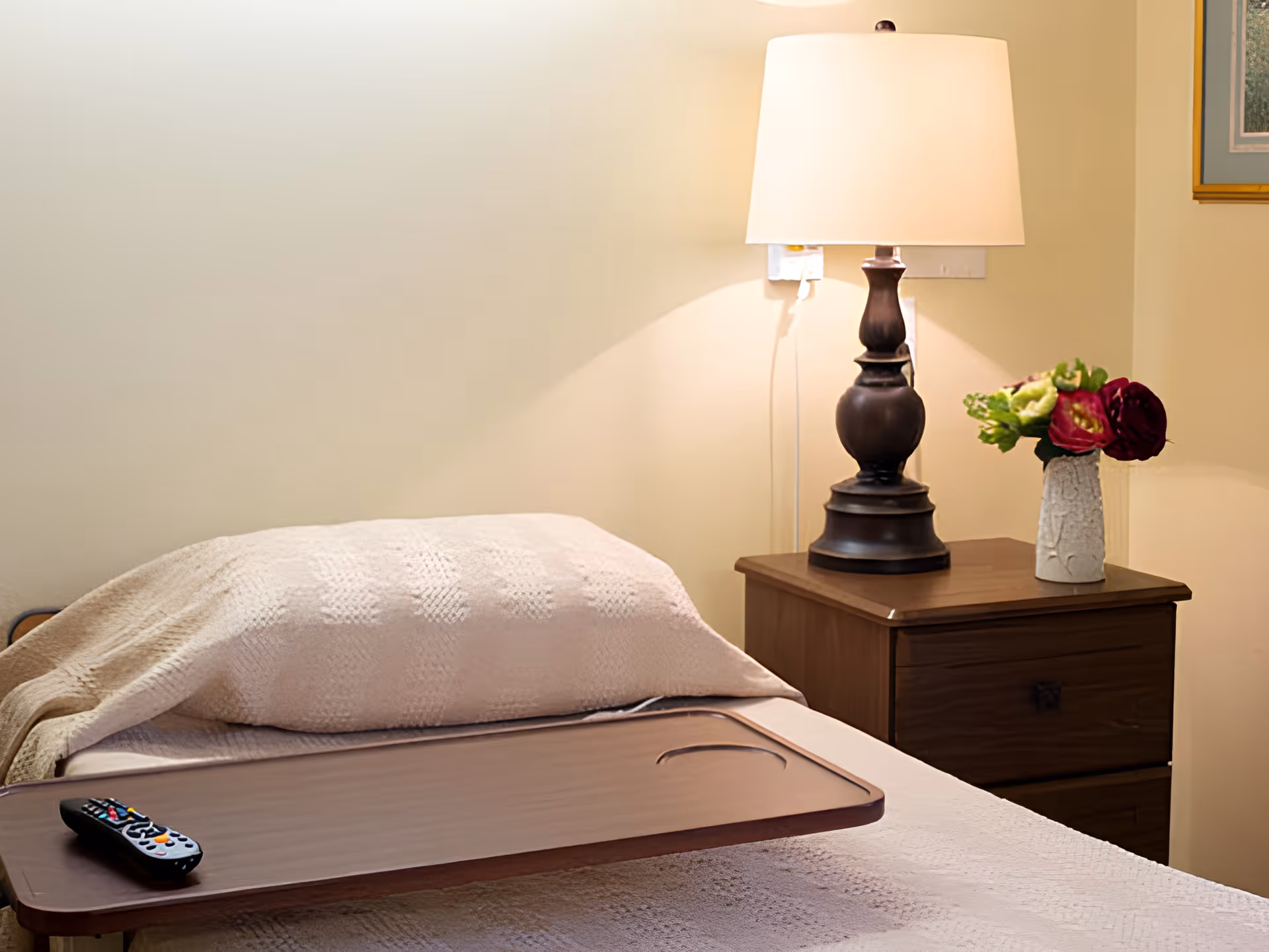 A neatly made bed with a beige textured blanket and a wooden tray holding a TV remote. Next to the bed is a wooden nightstand with a dark brown lamp and a white vase containing red and green flowers. The wall behind is light-colored with a framed picture partially visible.